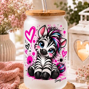 Könnte beinhalten: Ein mattierter Glastrinkbecher mit Bambusdeckel und klarem Strohhalm, bedruckt mit einem Zebra mit schwarz-weißen Streifen, blauen Augen sowie pinken und blauen Haarsträhnen, umgeben von pinken Herzen und Farbspritzern mit dem Schriftzug Sunny Louu.