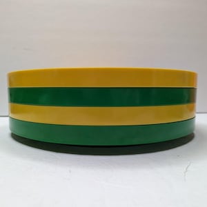 以下が含まれることがあります： 黄色と緑のストライプ模様の、4つの丸くて平らな物体が積み重なっています。物体は積み重ねられており、ストライプ模様を作り出しています。物体は固い素材でできているようです。直径は約20cmです。