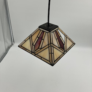 Puede incluir: Una lámpara colgante con un diseño geométrico. La pantalla es cuadrada con un patrón de vidrieras en crema, burdeos y blanco. La lámpara tiene un marco de metal negro y un cable negro.