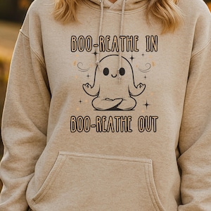 May include: A tan hoodie with a front pocket and drawstring hood. The front features a graphic of a meditating ghost with the text "BOO-REATHE IN" above and "BOO-REATHE OUT" below. The ghost is white with a smiling face.