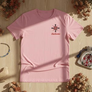 May include: A light pink t-shirt with a floral cross design and the name "Katherine" embroidered on the chest. The image includes dried flowers, a beaded necklace, a small bird figurine, and a drawstring bag with floral embroidery.
