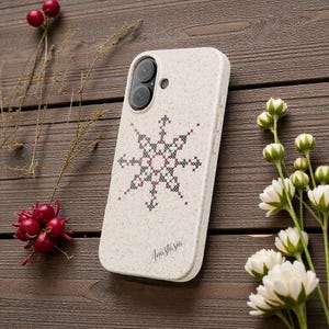 May include: A cream-coloured phone case featuring a cross-stitch snowflake pattern in black and red. The name "Anastasia" is printed in cursive. The case rests on a wooden surface, with floral and berry decorations.