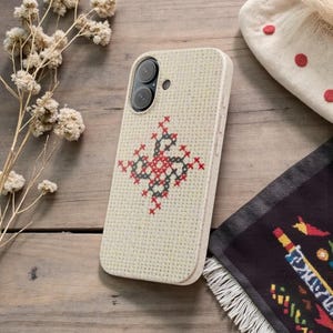 May include: A beige phone case with a cross-stitch design in red and black. The case has a textured appearance and a camera cutout. The phone case is on a wooden surface with dried flowers and a patterned textile.