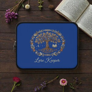Puede incluir: Una funda azul para portátil con una ilustración detallada de un árbol, un libro, una luna creciente y estrellas, rodeada por una corona. Las palabras "Lore Keeper" están escritas en oro debajo del diseño del árbol.
