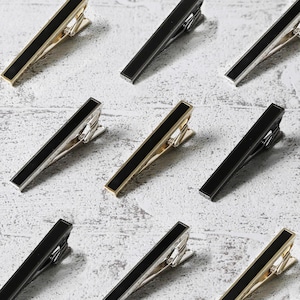 May include: A set of ten black and silver tie clips. The tie clips are made of metal and have a rectangular shape. The clips are arranged in a grid pattern on a white surface.