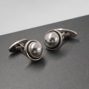 May include: A pair of silver cufflinks. Each cufflink features a large, polished, spherical center piece surrounded by a circular frame. The cufflinks have a classic design with a t-bar closure. The background is a dark gray surface.