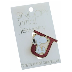 May include: Snoopy initial jewellery charm featuring Snoopy sitting inside a red letter U. The charm is on a white card with the text "Snoopy initial jewellery" and "United Feature Syndicate, Inc."