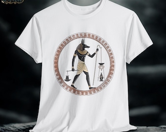 May include: White t-shirt featuring a circular design with an illustration of Anubis, the Egyptian god of the afterlife. The design includes a scale, a staff, and a lantern, all in a brown and gold color scheme. The shirt has short sleeves.