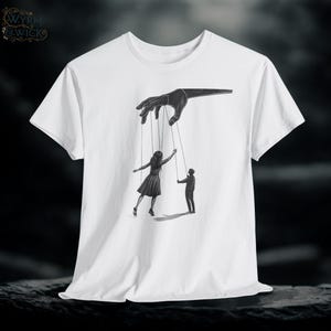 Può includere: T-shirt bianco con un'illustrazione in bianco e nero di una mano che manipola due figure con delle corde. Le figure sono una donna in abito e un uomo in giacca e cravatta. La maglietta ha un girocollo e maniche corte.