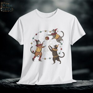 May include: White t-shirt featuring a whimsical illustration of three cats dressed as jesters, juggling a ball within a circle of colorful lights. The cats wear hats and costumes in red, yellow, and brown. The shirt has a classic crew neck.