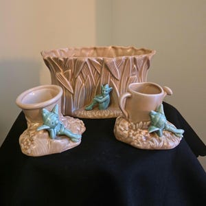 May include: A collection of vintage ceramic planters and vases in a light tan hue. The set features a large rectangular planter and two smaller vases, each decorated with a turquoise fairy figurine. The large planter has a textured, vertical design.