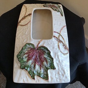 May include: Cream ceramic picture frame with a textured surface and a rectangular aperture. The frame is decorated with green and burgundy leaf designs and brown vine accents.