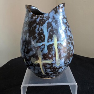May include: A ceramic vase with a dark brown base and blue and yellow splatter design. The vase has a unique shape with a wavy top and cross-like markings in blue and yellow. The vase is displayed on a clear plastic stand.