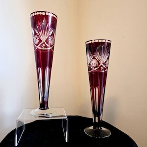 May include: Two tall, slender, ruby red glass vases featuring clear cut-glass patterns. Each vase has a flared rim with a circular motif, a body decorated with starburst and geometric engravings, and a clear glass base. The vases vary in height.