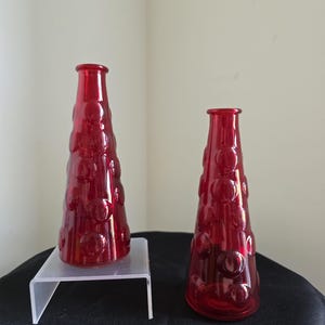 May include: Two red glass vases with a textured, bubble-like design. The vases are cone-shaped, with a narrow neck and a wider base. One vase sits on a clear acrylic stand, while the other rests on a black surface. The background is a neutral colour.