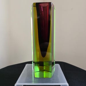 May include: A rectangular glass vase with a vibrant colour scheme. The vase features a gradient of colours, including green, yellow, and a deep red-purple. The geometric design and colour transitions create a striking visual effect. The vase is displayed on a clear acrylic stand.