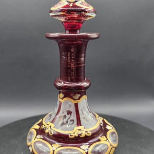 May include: A ruby red glass perfume bottle with a decorative stopper. The bottle features gold and white painted floral designs and oval accents. The stopper is also red with gold and white accents.