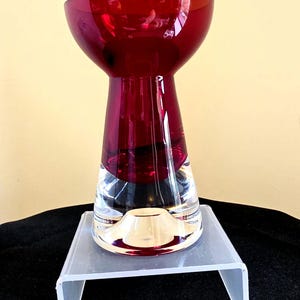 May include: A striking ruby red glass vase with a clear base. The vase features a wide, rounded top that narrows to a neck, then flares out at the base. The clear base provides a contrast to the rich red colour.