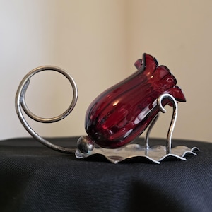 May include: A ruby red glass bud vase in a tulip shape, set in a silver-toned metal holder. The holder has a decorative, scalloped base and a curved, spiral accent. The vase is on a black surface.