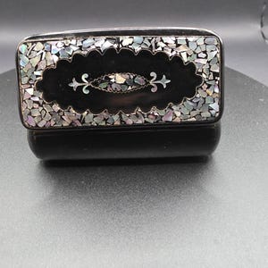 May include: A rectangular black box with a lid, featuring an inlaid design of iridescent shell fragments. The lid's centre has a dark oval with a decorative floral design. The box is likely used for jewellery or small items.
