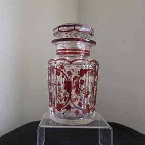 May include: A clear glass jar with a matching lid, featuring vertical fluted panels and a deep red floral pattern overlay. The jar has a cylindrical body with a slightly tapered neck and a faceted stopper, showcasing intricate red botanical details.