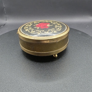May include: A small, round antique jewellery box. The black lid has a red floral design and gold filigree. The box has a gold-coloured metal base and three small feet. The lid is hinged. The box appears to be in good condition.