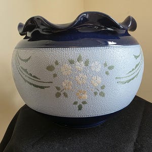 May include: A round ceramic bowl with a textured pale blue exterior, decorated with floral and leaf designs in muted green and yellow. The rim and base are a deep navy blue, with a ruffled edge. A decorative piece.