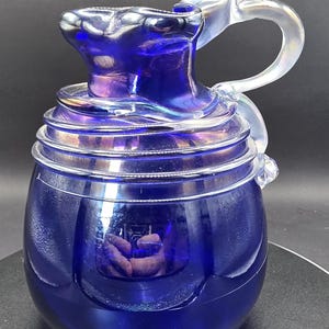 May include: A deep blue glass jug with a clear, curved handle and spout. The jug has a rounded base and horizontal ridges around the middle. The handle is shaped like a stylized bird or wing.