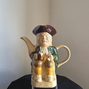 May include: A ceramic teapot in the form of a seated man, wearing a brown hat and green coat. The teapot has a tan body, a spout, and a handle. The man holds two brown kegs. The teapot is on a black surface.