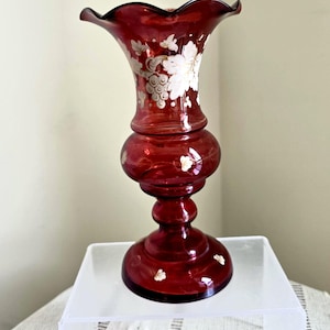 May include: A deep red glass vase with a wavy rim and a rounded body. The vase is decorated with white floral and grape motifs. The base is circular. The vase is displayed on a clear acrylic surface.