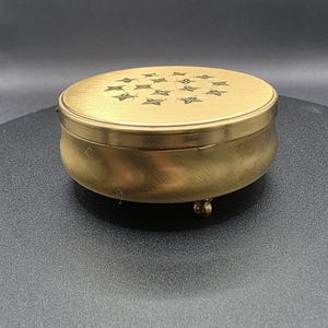 May include: A small, round, antique brass trinket box with a lid. The lid features a pattern of star-shaped designs. The box has a brushed finish and stands on three small, ball-shaped feet. The background is dark.