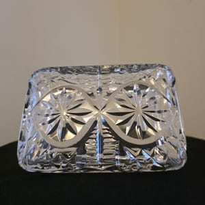 May include: A clear, rectangular crystal dish with a starburst and diamond pattern. The dish has a lid and is likely used for serving or storage. The crystal reflects light, creating a sparkling effect. The design is symmetrical and elegant.