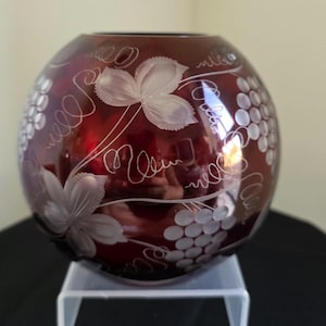 May include: A spherical, ruby-red glass vase featuring etched grape and leaf patterns. The clear, detailed carvings contrast with the coloured glass. The vase is a decorative item, suitable for home decor.
