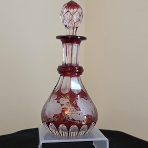 May include: A ruby red glass perfume bottle with a decorative stopper. The bottle features etched floral designs and a bulbous shape. The stopper is a matching red with a detailed, geometric pattern. The bottle is displayed on a clear stand.
