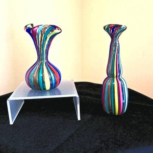 May include: Two glass vases featuring vertical stripes in vibrant colours including blue, red, yellow, and teal. One vase has a rounded bulbous base with a flared rim, while the other is a taller, slender vessel with a narrow neck and cylindrical body.