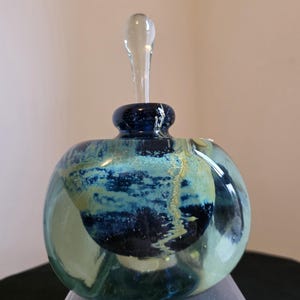 May include: A decorative glass perfume bottle with a clear glass stopper. The bottle is round with a dark blue neck and a body featuring blue, green, and yellow swirls. The bottle sits on a clear plastic stand.