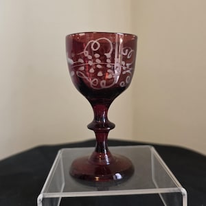 May include: A deep red glass goblet featuring a delicate, etched white pattern of swirling lines and small dots around the upper bowl. The vessel has a slender stem with a central decorative node and a wide, circular base for stability.