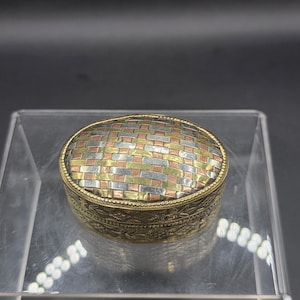 May include: An oval metal box featuring a woven pattern on the lid in alternating strips of brass, copper, and silver-toned metal. The sides of the box are decorated with an engraved geometric pattern and a beaded border along the top edge.