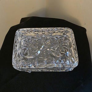 May include: A clear, rectangular glass dish with a detailed starburst and geometric pattern. The dish has a slightly raised edge and is likely used for serving or display. The glass reflects light, creating a sparkling effect.