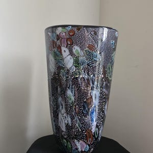 May include: A tall, cylindrical glass vase featuring a dark, speckled black base embedded with numerous multicoloured glass inclusions in shades of blue, green, brown, and white, creating a textured, confetti-like appearance throughout the surface.