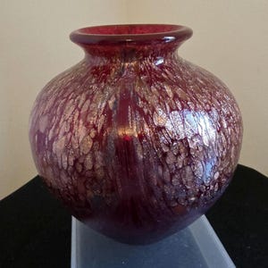 May include: A round, burgundy glass vase with a wide body and a narrow neck. The vase has a speckled, metallic gold pattern throughout. The vase is on a clear plastic stand. The vase is approximately 20 cm in height.