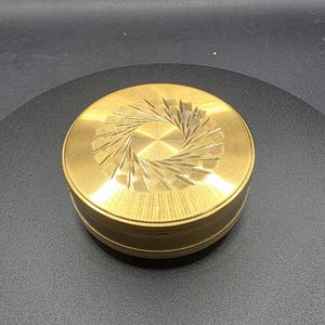 May include: A circular, gold-toned metal box with an engraved lid design. The lid features a central starburst pattern with radiating lines. The box is displayed on a dark surface. The box is approximately 7 cm in diameter.