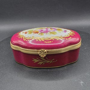 May include: An oval trinket box in a deep pink colour. The lid is decorated with a floral design featuring pink and purple flowers on a white background. Gold-coloured trim and a decorative clasp enhance the box's elegance. Likely made of porcelain.