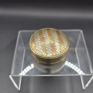 May include: A round metal trinket box featuring a lid with a woven pattern of alternating copper, brass, and silver-toned rectangular strips. The sides of the box are a solid, muted gold-toned metal, creating a compact and circular container.