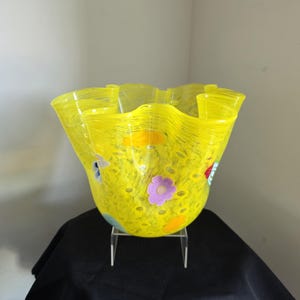 May include: A bright yellow glass bowl with a ruffled, wavy rim. The translucent yellow glass features a textured surface with embedded colourful floral accents in shades of purple, orange, and blue, creating a vibrant, multi-layered appearance.