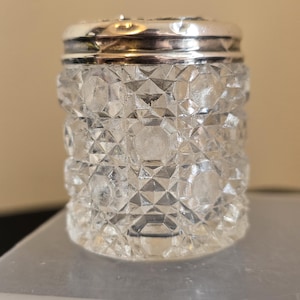 May include: A clear, faceted glass jar with a silver-coloured lid. The jar has a cylindrical shape with a diamond-like pattern. The lid is shiny and reflects light. The jar is likely for storing small items or spices.