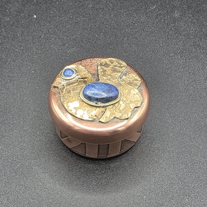 May include: A small, round copper box with a brass lid featuring a floral design. The lid is adorned with two blue lapis lazuli stones. The box has a decorative, ribbed pattern around the sides. The box is likely used for storing small items.