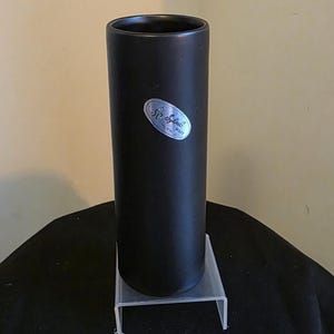 May include: A tall, cylindrical black vase with a small, oval label. The vase is displayed on a clear, rectangular stand. The label appears to read "SP Sylvate Ware".
