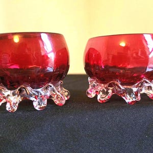 May include: Two ruby red glass bowls with clear, textured bases. The bowls have a rounded shape and sit on a clear, decorative base. The glass has a glossy finish, reflecting light. These decorative bowls are ideal for home decor.