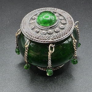 May include: A small, round, dark green glass jar with a silver-coloured lid. The lid features a large green gem in the centre and is decorated with intricate patterns. Small chains with green beads hang from the jar.